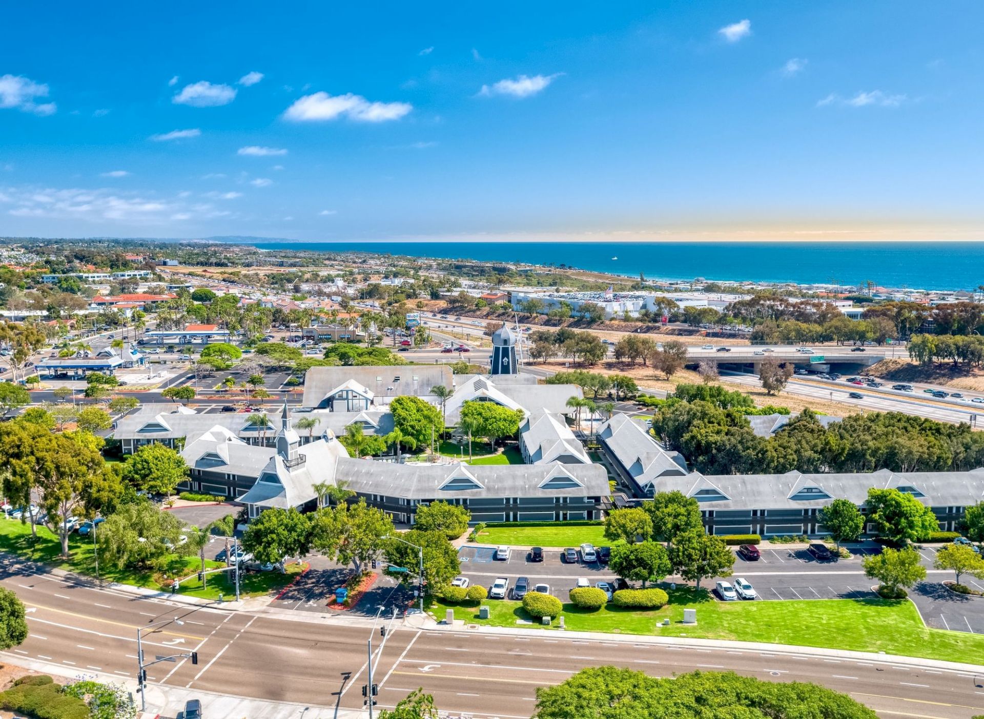 Aerial view of a coastal town: buildings and highways in front, blue ocean on the horizon under a sunny sky.