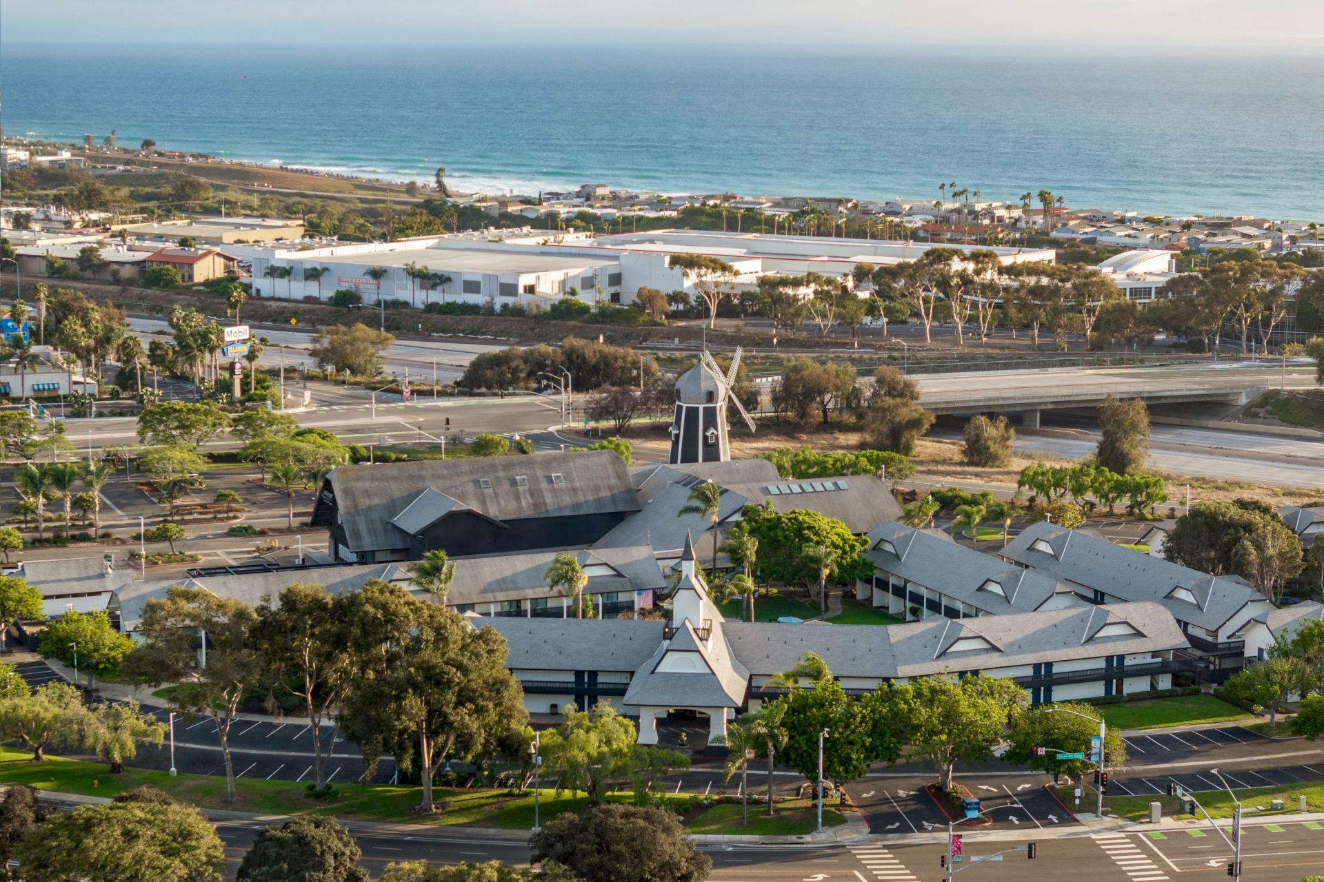 Coastal town view with palm trees, a windmill-shaped building, roads, and a long beach along the blue ocean.