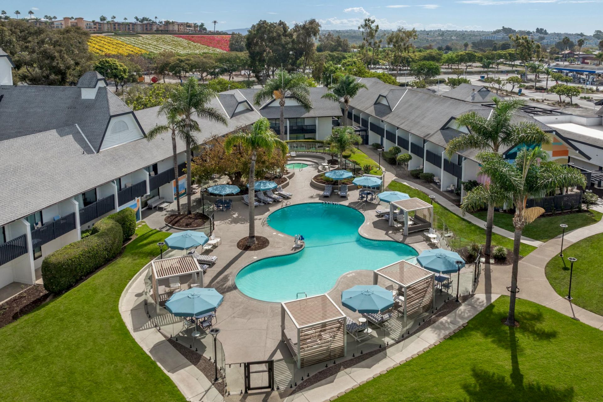 A sunny hotel courtyard with two turquoise swimming pools, lounge chairs, umbrellas, palm trees, and low-rise guest buildings.