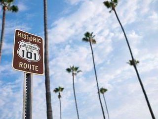 A &ldquo;Historic Route 101&rdquo; sign stands among tall palm trees against a blue, partly cloudy sky.