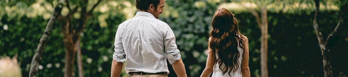 A couple walks hand-in-hand along a path under leafy trees, holding drinks in a sunny, romantic park setting.