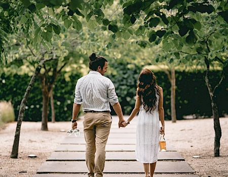 A couple walks hand-in-hand along a path under leafy trees, holding drinks in a sunny, romantic park setting.
