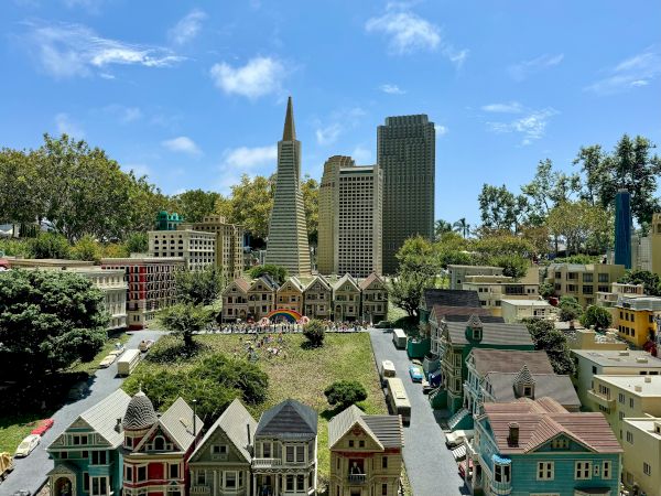 A sunny city skyline of tall office towers over colorful historic-style houses and trees in a park-like courtyard.