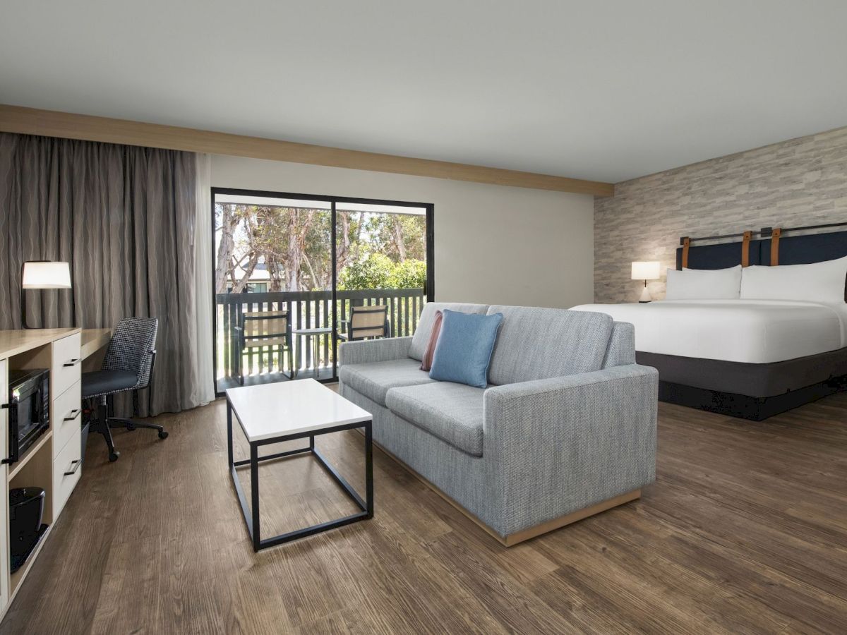 A modern hotel suite with a king bed, gray sofa, coffee table, lamps, and sliding doors to a balcony with trees outside.