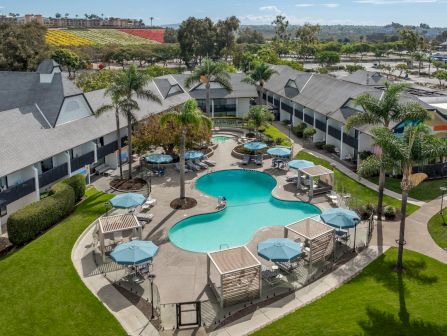A sunny hotel courtyard with two turquoise swimming pools, lounge chairs, manicured lawns and palms, plus nearby farm-style crops in view.