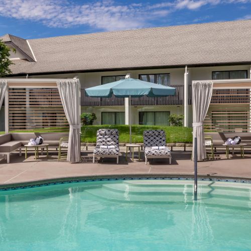 A sunny backyard patio with a swimming pool, comfy lounge chairs, and a shaded gazebo-style seating area with white curtains.
