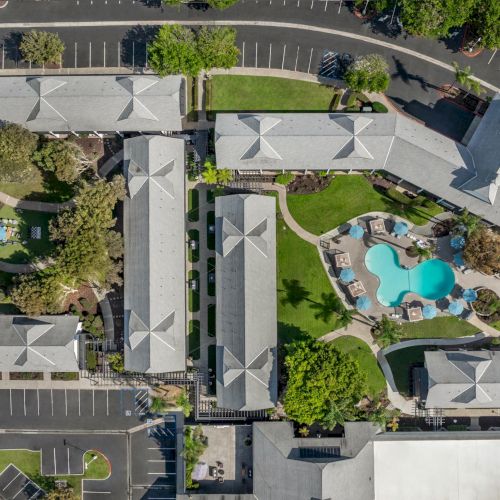 Aerial view of a building complex with gray roofs, landscaped grounds, a court area, and a turquoise swimming pool.