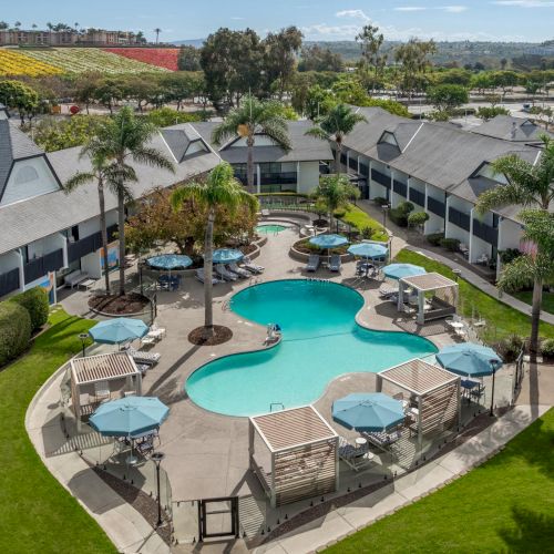 A sunny resort courtyard with two turquoise pools, lounge umbrellas, and apartment buildings, overlooking green hills and vineyards.
