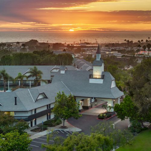 Sunset over the ocean behind a palm-filled resort, with rooftops and warm light glowing over the coastline.