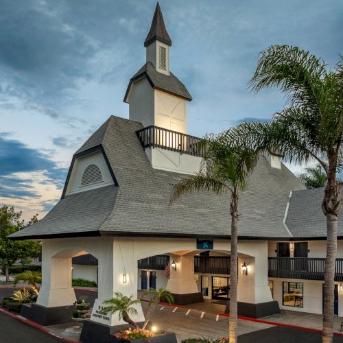 A white chapel with a tall steeple and lit entrance, surrounded by palm trees under a cloudy evening sky.