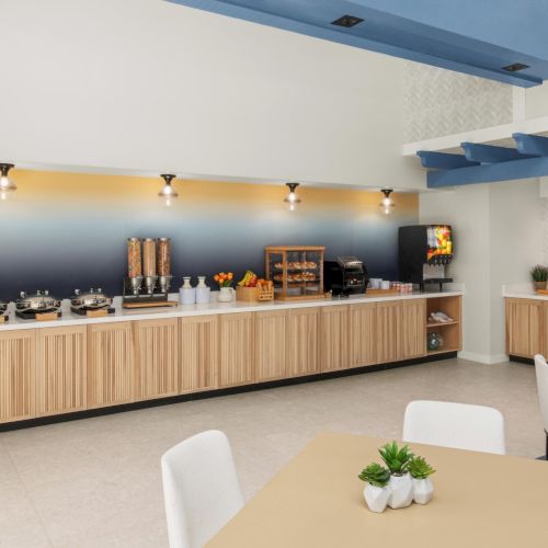 An open buffet serving area with countertop dispensers, coffee/tea setup, and grills, plus seating in a modern lounge.