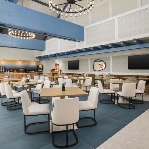A bright hotel breakfast buffet area with tables, chairs, a snack counter, and overhead lights under blue beams.