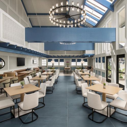 A bright modern lounge dining area with rows of tables and chairs, paneled walls, and a large chandelier overhead.