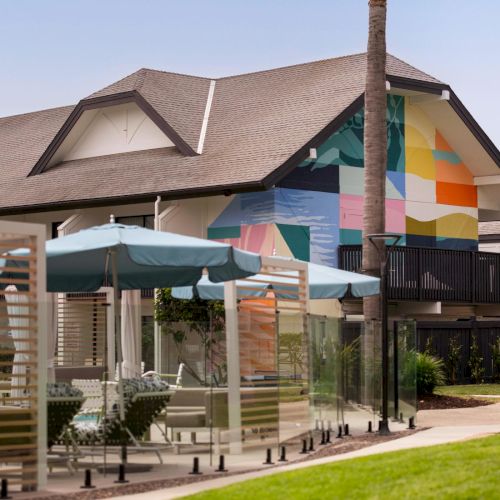 Outdoor patio by a house with a colorful mural on the wall, umbrellas, and a tall chimney rising above the roof.