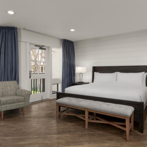 A modern bedroom with a bed, bedside lamps, an armchair, and blue curtains beside large windows, lit by recessed ceiling lights.