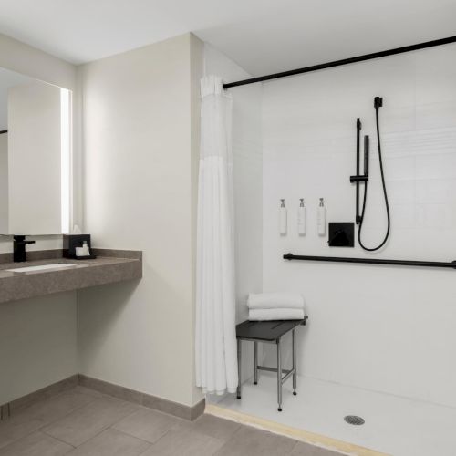 A modern bathroom with a wall sink and countertop, and a rainfall shower with black fixtures, bottles, and neatly folded towels.