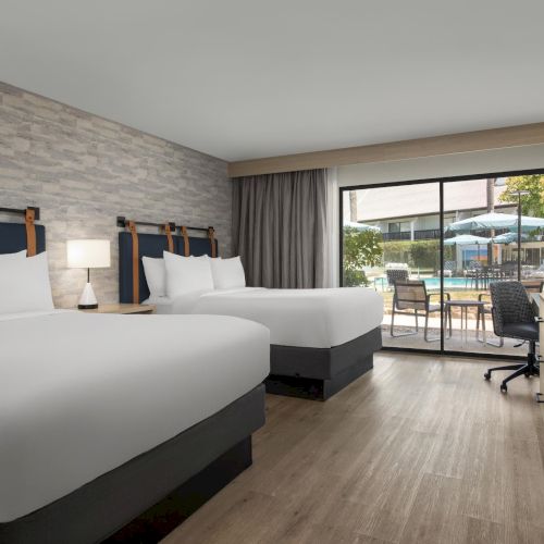 A modern hotel room with twin beds, desk and TV, and a view through glass doors to an outdoor pool patio.