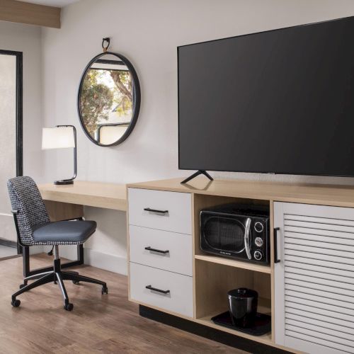 A modern living room shows a flat-screen TV on a wooden console with an office chair, oval mirror, and large windows.