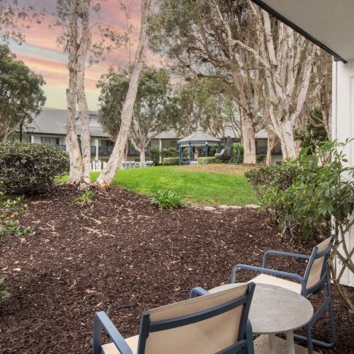 An outdoor courtyard with tall trees, trimmed grass, a small blue gazebo, and patio chairs by a building in dusk light.