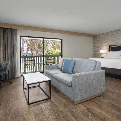 A modern hotel room with a gray sofa, neatly made bed, side lamps, and a large sliding door to a balcony with trees outside.