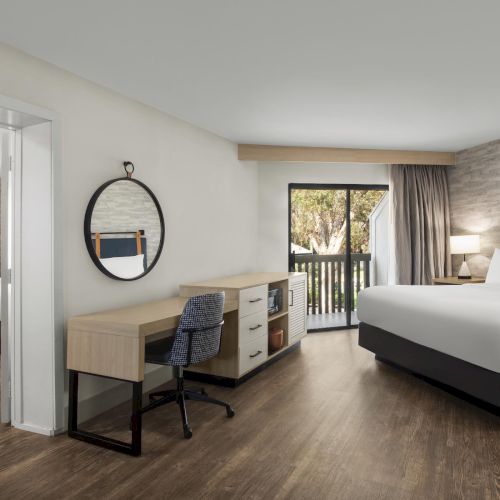 A modern hotel room with two beds, a desk, round mirror, and large patio doors with curtains.