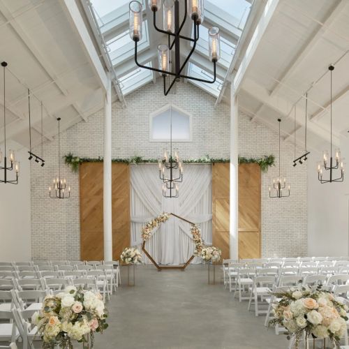 A bright wedding venue with rows of white chairs, floral decor, and lit chandeliers hanging above the ceremony arch.