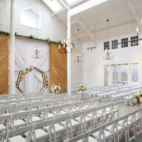 Bright wedding chapel with rows of white chairs, a floral arch, and hanging lantern lights under a vaulted ceiling.