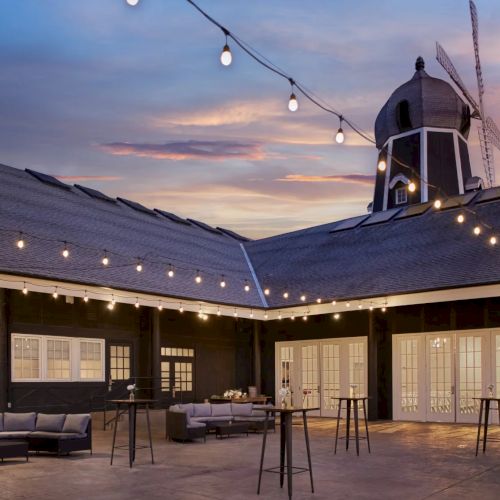 A cozy courtyard at dusk with string lights, patio furniture, and a towering windmill-like structure under a pink sky.